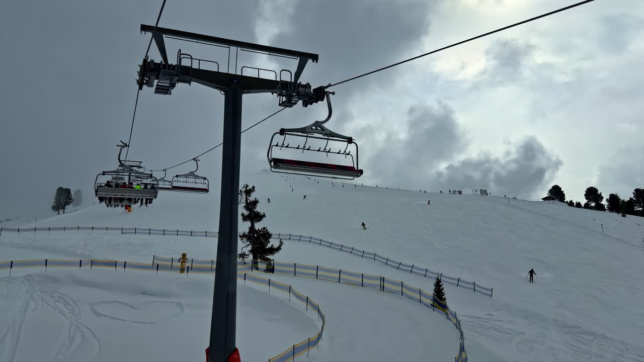 Video POV di una seggiovia che trasporta sciatori su piste da sci battute con recinzioni di sicurezza e sciatori che scendono sullo sfondo presso la stazione sciistica di Nauders, Austria