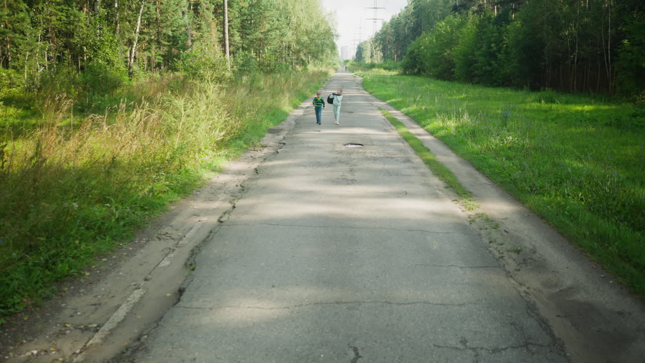 Distant rear view of two children walking side by side on long tarred road surrounded by forest trees and greenery, heading home under daylight with calm mood and natural shadows on rural path