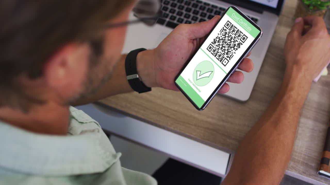 Man at desk holding smartphone with covid vaccination certificate and qr code on screen