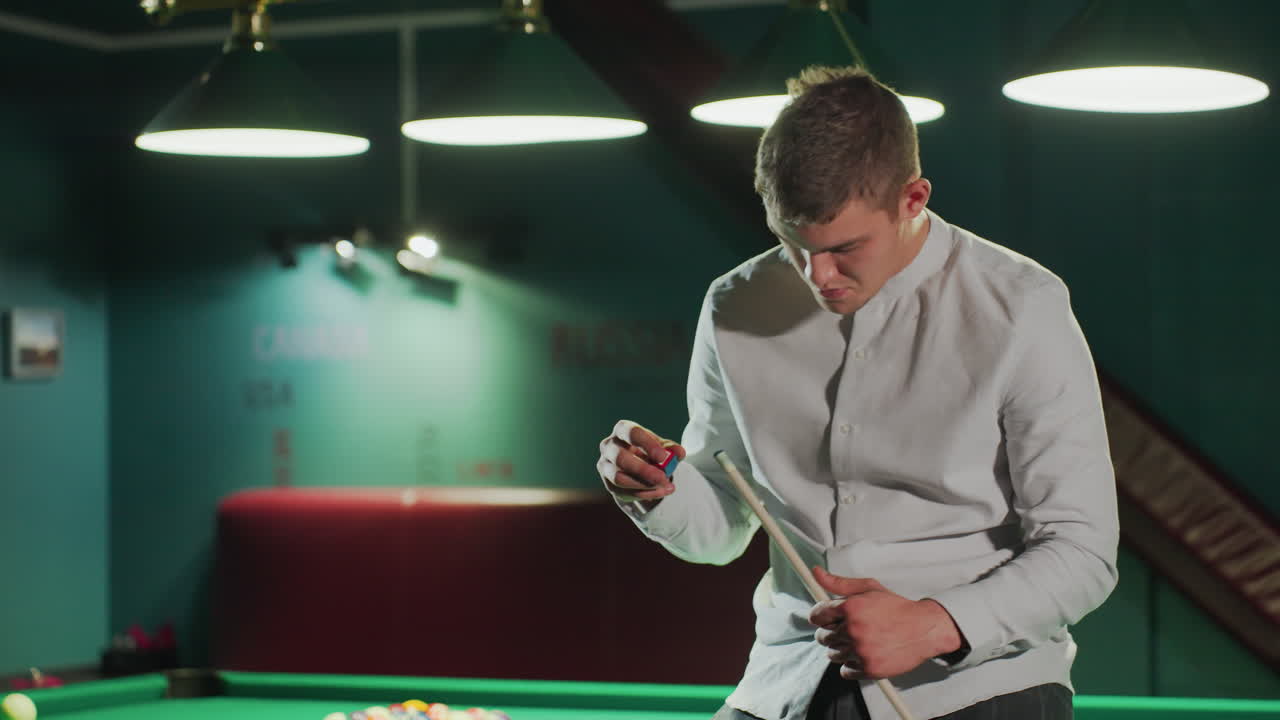 primer plano de un joven con camisa blanca y pantalones grises sentado en una mesa de billar verde, sosteniendo un palo de taco y aplicando cuidadosamente la cubierta roja con una expresión enfocada mientras la cámara hace zoom en la luz tenue
