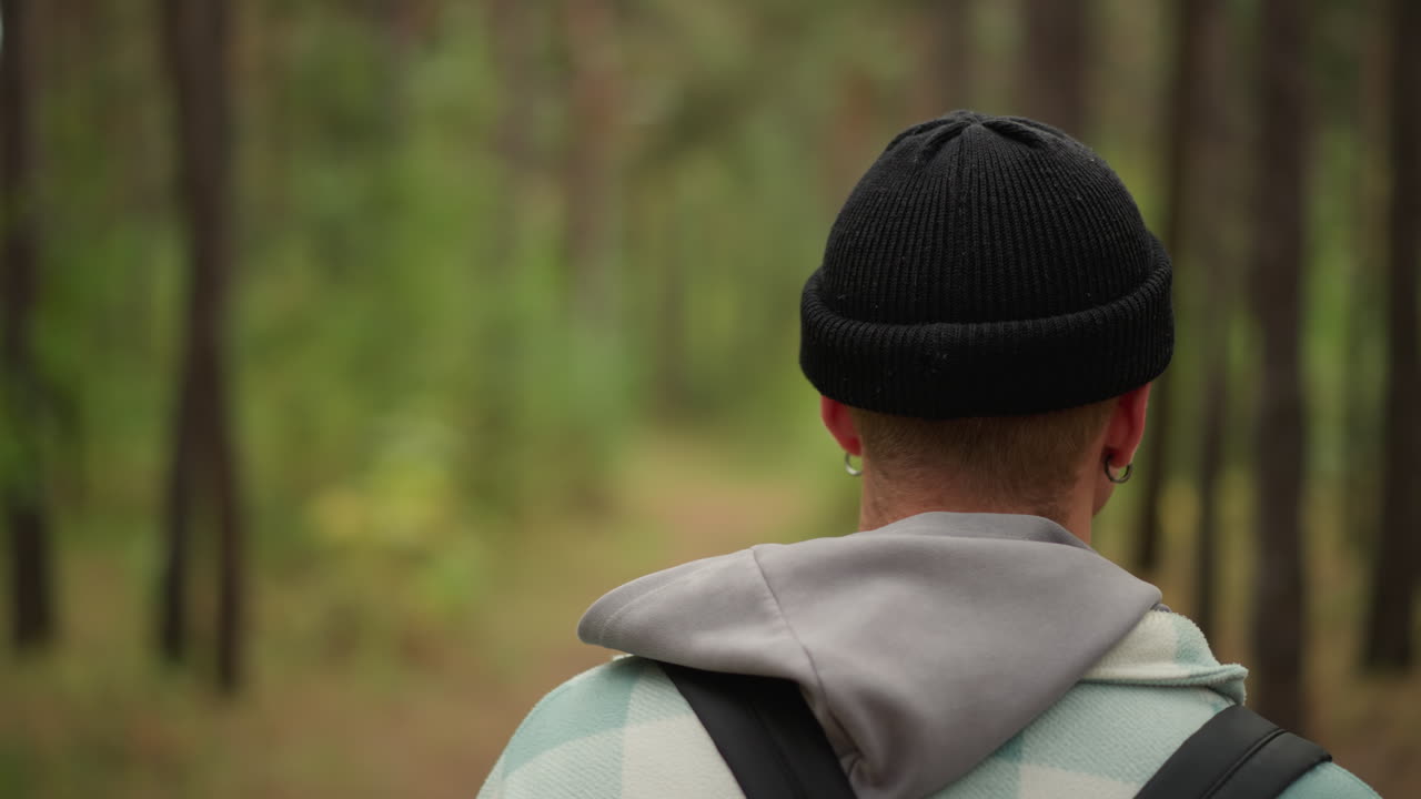 close portrait of man turning head in pine forest, checking beanie and scanning surroundings with contemplative expression, subtle earring detail and backpack strap visible, soft ambient light