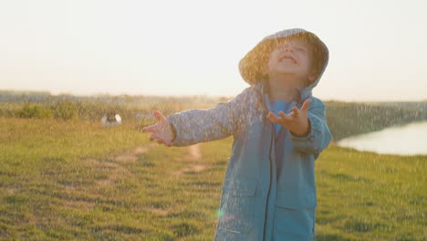 kid holds palms under warm spring rain in field cheerful toddler boy wearing raincoat has fun at