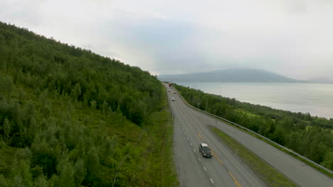 cars traveling on norwegian highway with green slope forest next to fjord