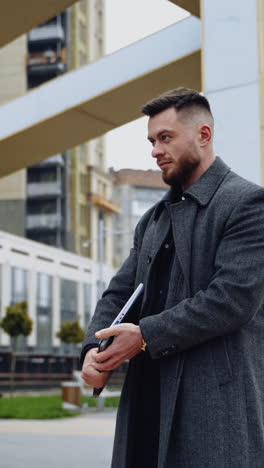 businessman with a laptop outdoors. handsome man in a coat working on wireless computer in the city street. business concept. vertical video