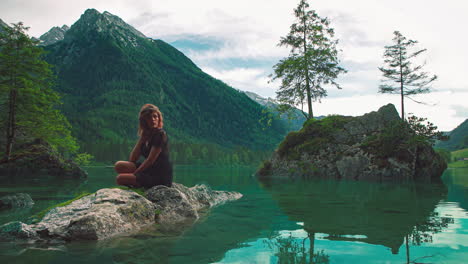 in this 4k uhd cinemagraph a young brunette woman is sitting on a rock at the scenic mountain lake hintersee in bavaria, a perfect travel destination in germany with the water moving gently