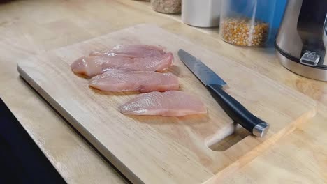 Tiro-Deslizante-De-Trozos-De-Pechuga-De-Pollo-Y-Un-Cuchillo-De-Chef-En-Una-Tabla-De-Cortar-De-Madera-En-Una-Cocina-Casera