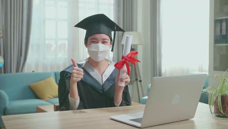 excited asian woman wearing protection face mask, thumb up and showing off a university certificate to camera. pretty female graduate wearing a graduation gown and cap sitting on the living room