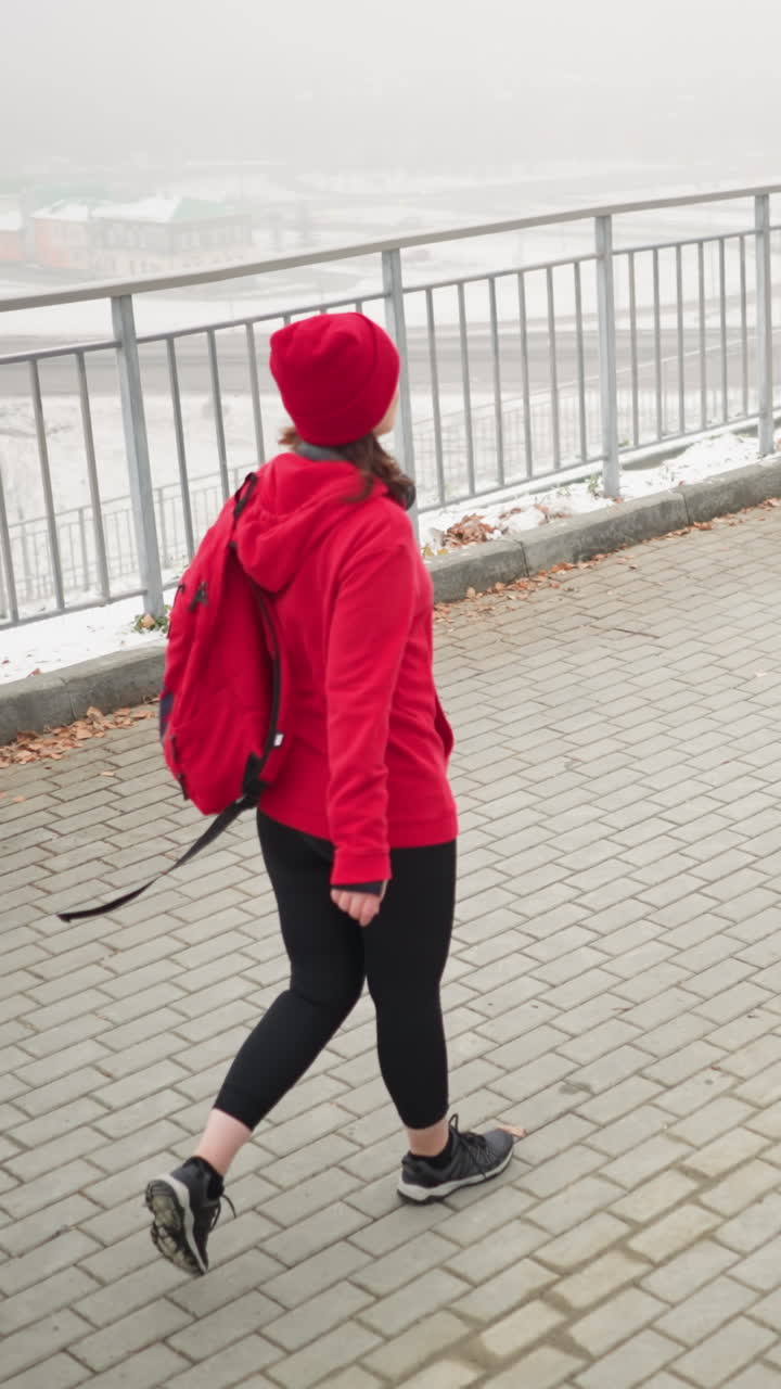 Back view of woman walking on interlocked pathway with bag over shoulder near iron railing overlooking foggy urban scene with cars below bridge and blurred office buildings in background