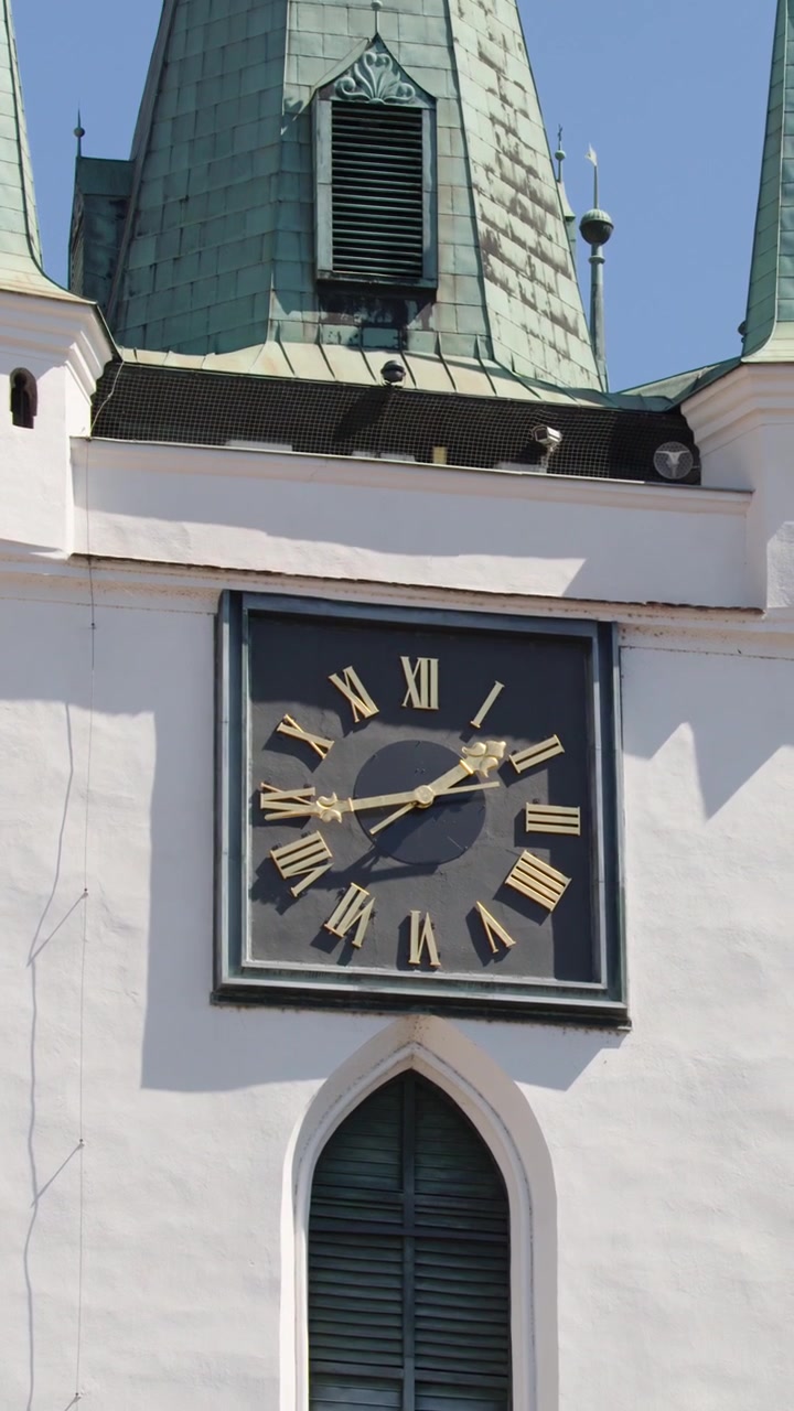 Camera tilts upward past ornate clock and windows to green spires against clear blue sky