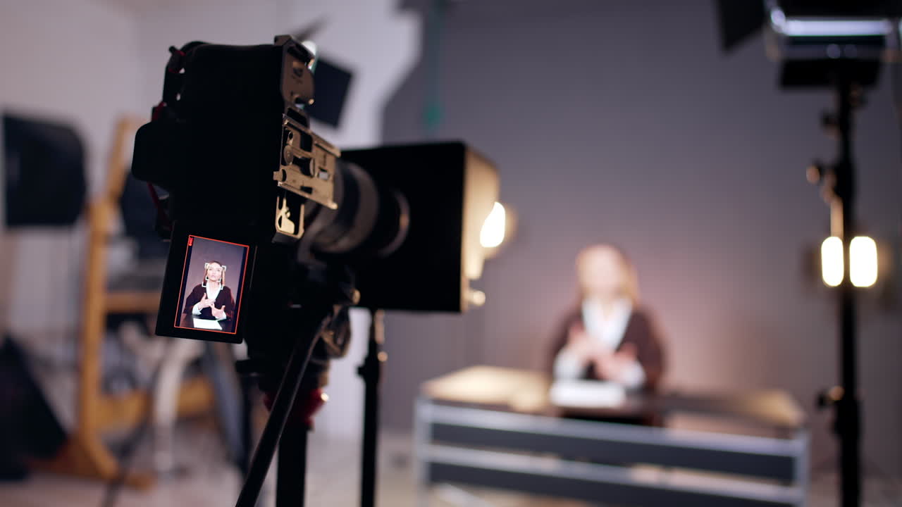 Professional camera set on tripod filming a lady in photo studio. Display shows the female narrator. Blurred backdrop.