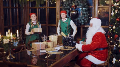elves in green suits and red hats is dancing with gifts in their hands and putting it on the table with letters of congratulations and a magnifying glass near santa claus on the background of the christmas tree and new year decorations in the room