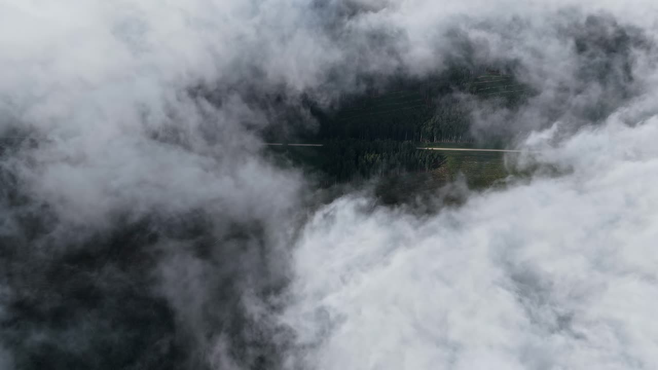 A hidden road cuts through a dense forest, partially revealed beneath thick mountain clouds. Europe
