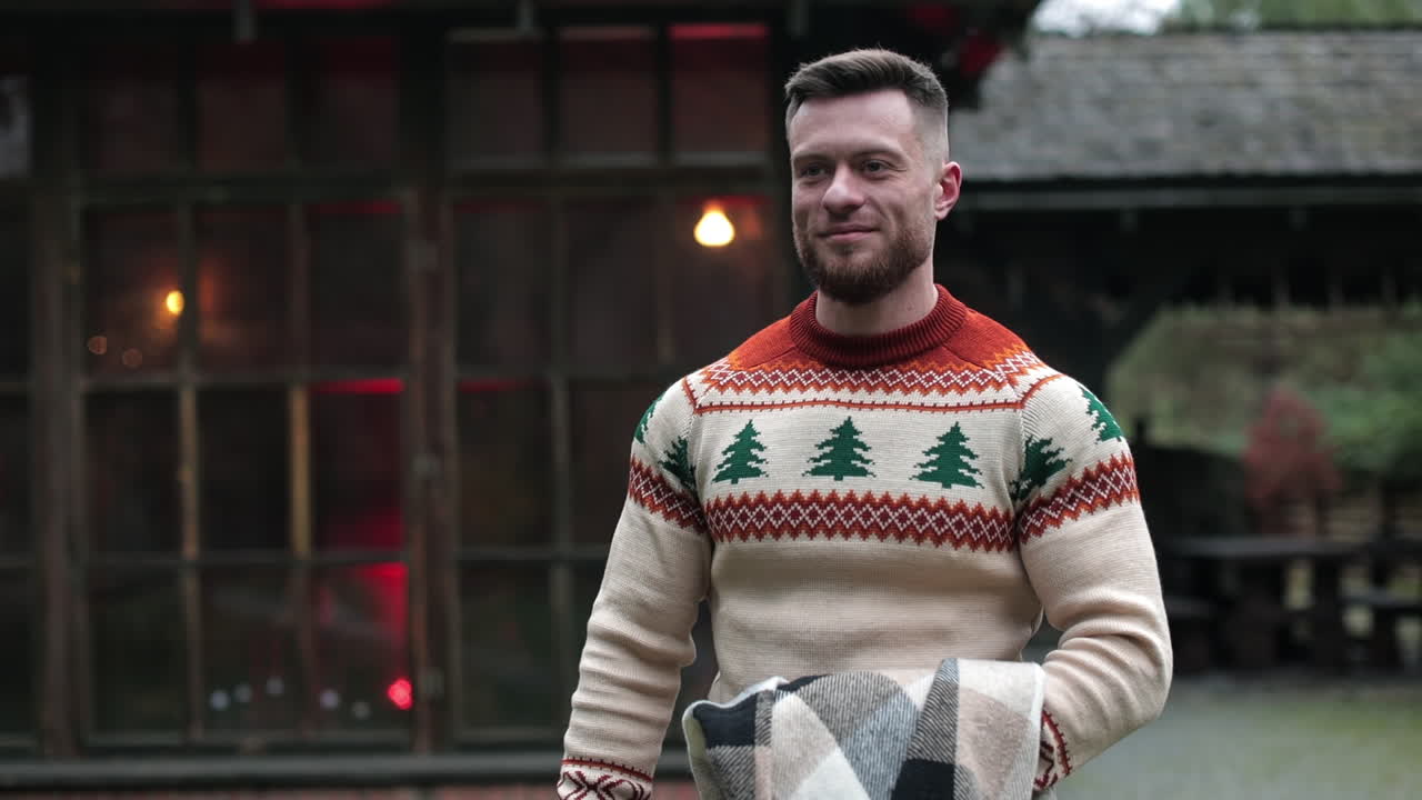 Smiling young man walks somewhere carrying warm blanket on his hand. Bearded male outside the big old rural house. Rural mansion with lights inside at the background.
