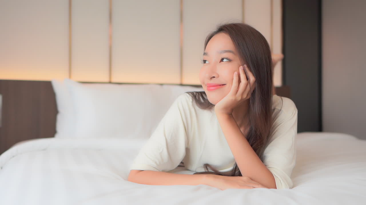 크고 편안한 침대에 누워 있는 예쁜 젊은 여성이 손에 턱을 기대고 미소를 짓고 있다