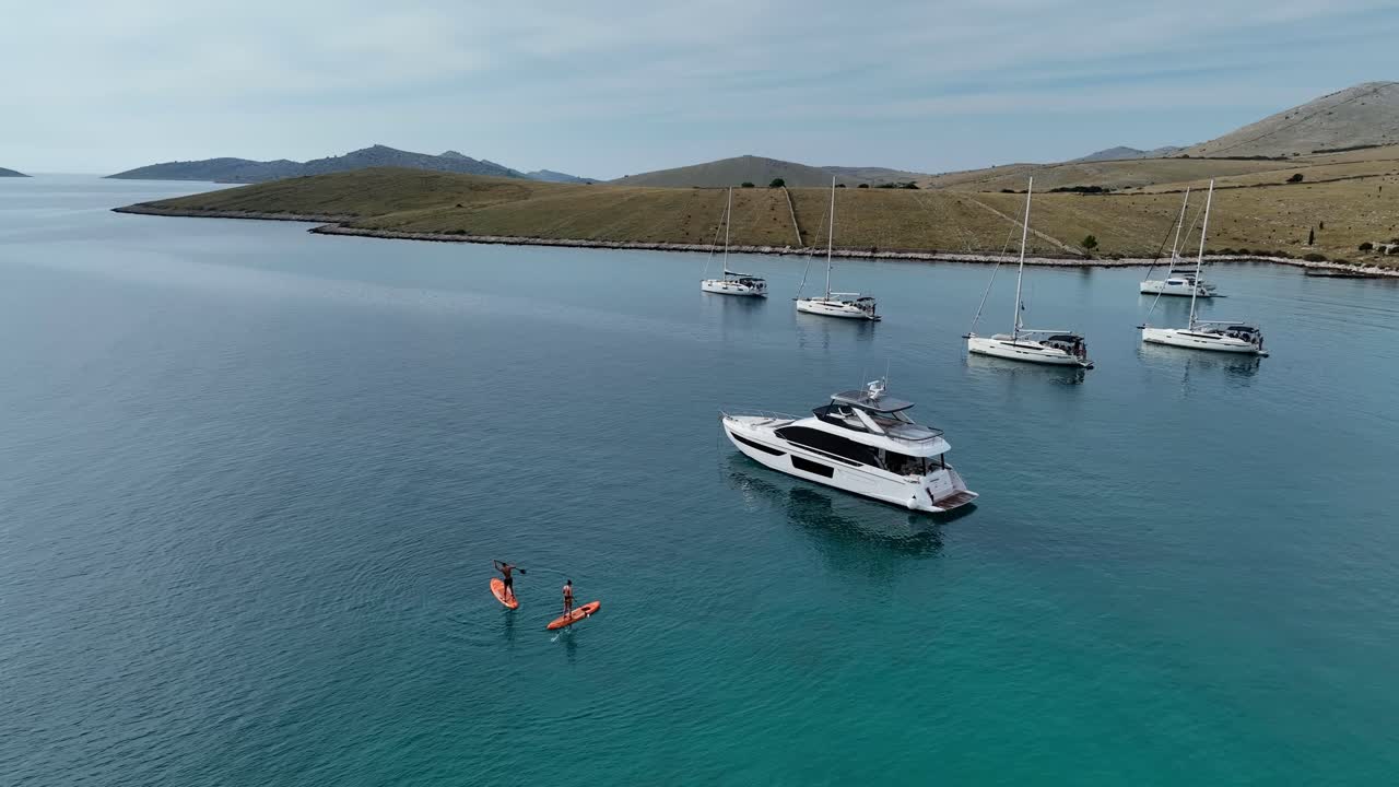 Sailboats yachts anchored in a bay with clear waters and surrounding hills in Kornati, Croatia as friends paddleboard in water