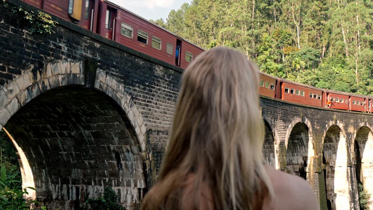 A young woman stands at a viewpoint near the Nine Arch Bridge in Ella, Sri Lanka, gazing at a colorful train as it moves across the historic stone arches.