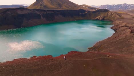 Aerial-shot-of-Crater-with-blue-water-in-it-,-couple-standing-on-edge,-mountains-in-the-background