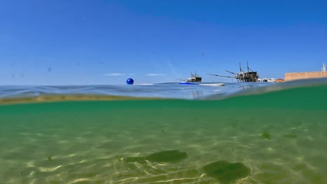 Vista-Medio-Subacuática-Inusual-De-Dos-Raquetas-De-Tenis-De-Playa-Y-Bola-Azul-Flotando-En-La-Superficie-Del-Agua-Del-Mar-Con-Plataformas-De-Madera-De-Pesca-Trabocchi-En-Segundo-Plano