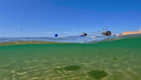 vista medio subacuática inusual de dos raquetas de tenis de playa y bola azul flotando en la superficie del agua del mar con plataformas de madera de pesca trabocchi en segundo plano