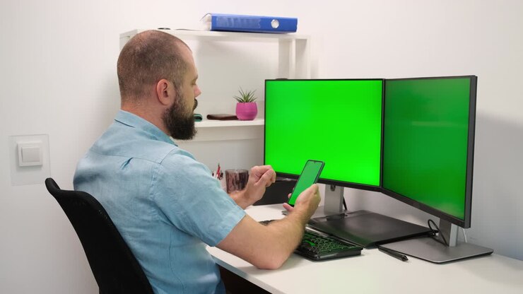 Uomo d'affari che lavora e digita su telefono cellulare e computer con due monitor, svolgendo un lavoro multitasking su schermo verde.