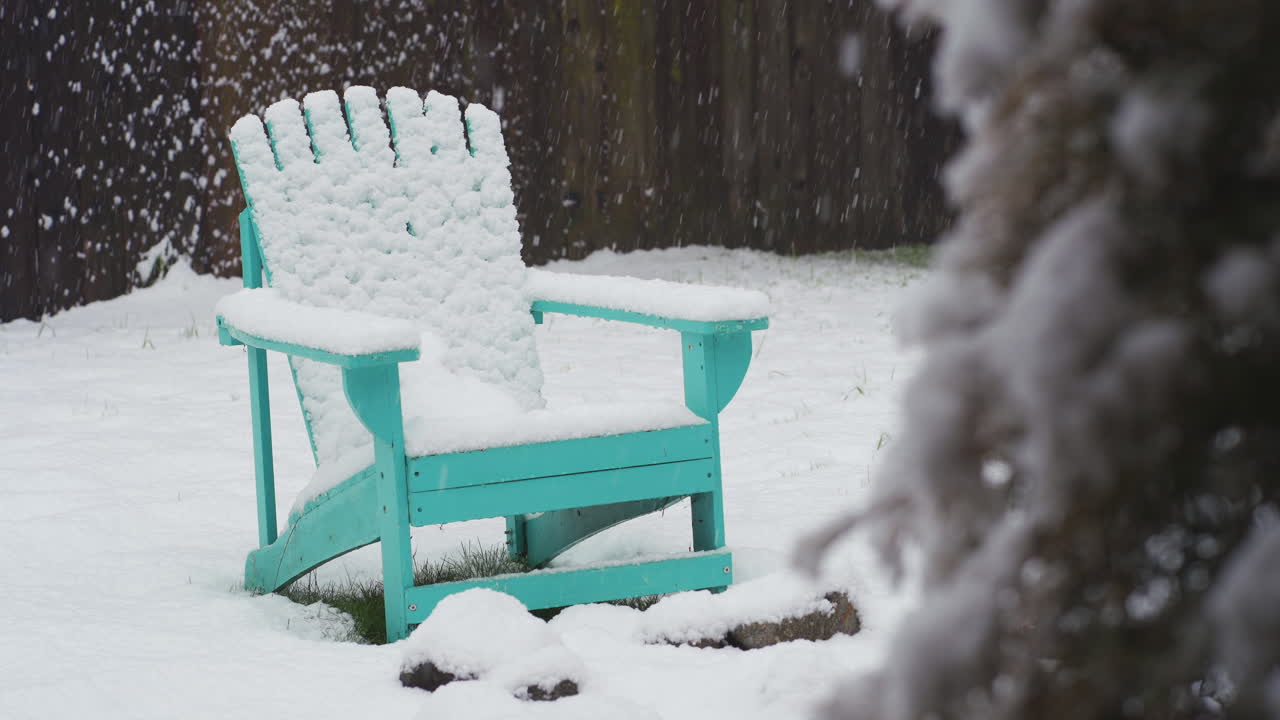 foto fija de nieve cayendo en un patio cubierto de nieve sobre una silla de jardín verde azulado frente a una valla de madera.