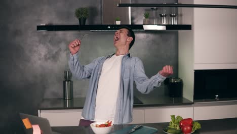 joven súper emocionado sosteniendo la cuchara como un micrófono cantando canciones y bailando mientras cocina en la cocina, hombre despreocupado preparando