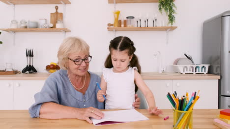 a concentrated little girl drawing sitting at table in kitchen with her happy grandma and mom while all together having a video call and looking at camera