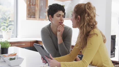 Happy-caucasian-lesbian-couple-standing-at-counter,-talking-and-using-tablet-in-sunny-kitchen