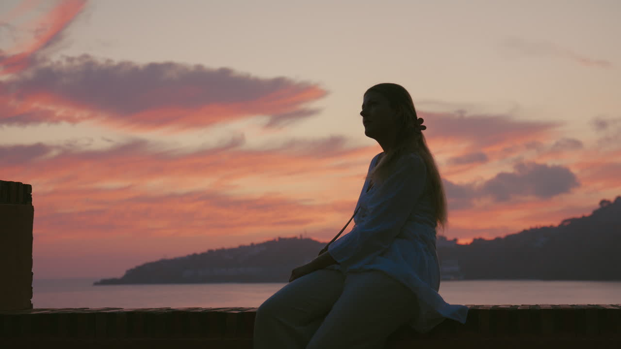 Wide side profile silhouette of a woman seated calmly on a terrace ledge, framed by dramatic pink and orange clouds. Stillness and quiet awe