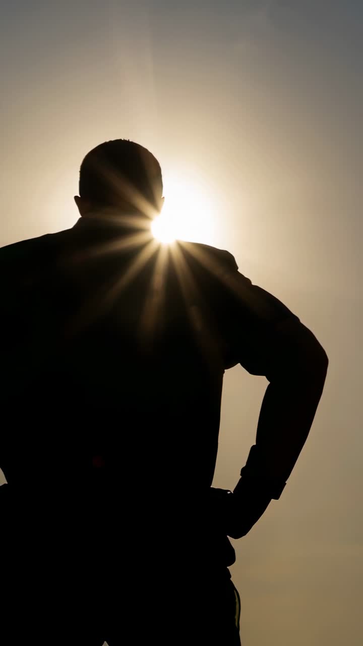 Vídeo vertical: Homem de camiseta em pé, com as mãos na cintura, observando o pôr do sol, com o relógio capturando o brilho solar em movimento.