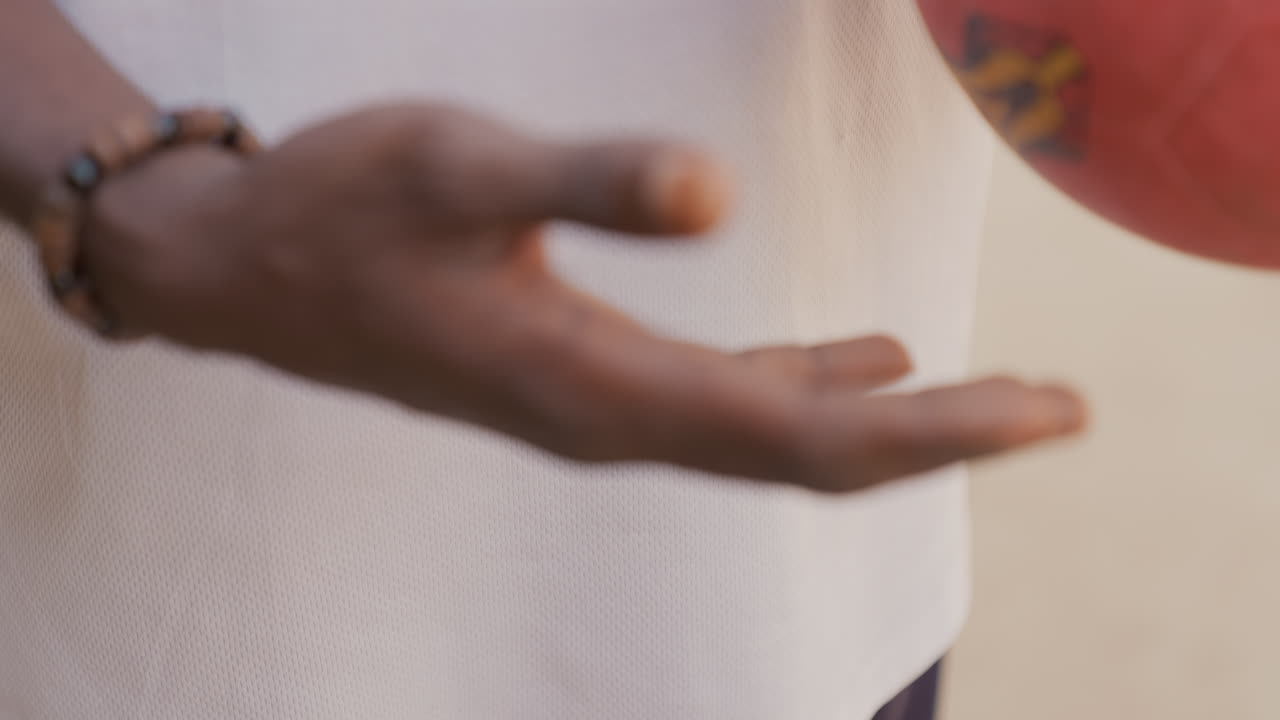 Black hands holding worn ball, closeup dusty red soccer ball cradled by black male player in white jersey, sandy fingertips and bracelet, slowmotion practice on dirt pitch, intimate preparation
