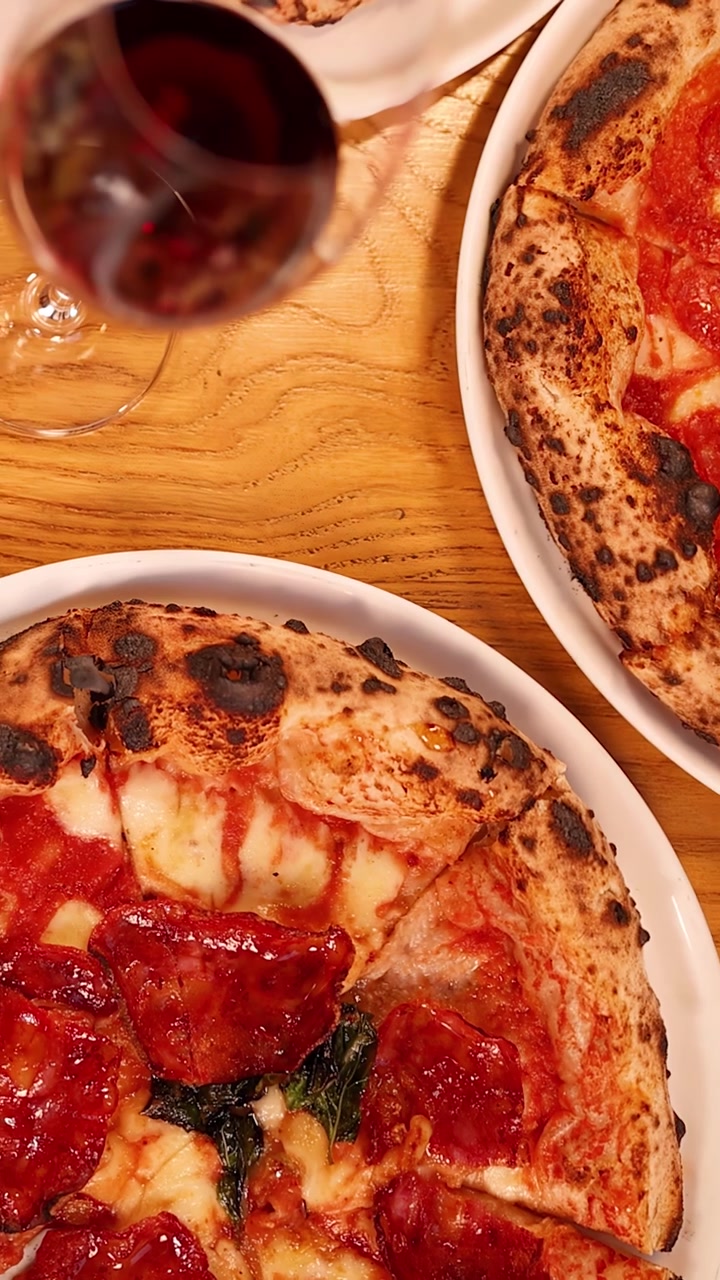 Movimento della telecamera dall'alto verso il basso che rivela diverse pizze al salame piccante e bicchieri di vino rosso su un tavolo di legno.