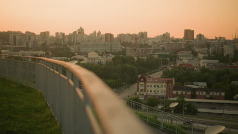 urban cityscape at sunset with car driving down street, iron railing in foreground leading viewer's eye towards skyline, lush greenery visible between buildings