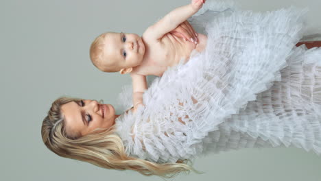 beautiful blonde long-haired woman with a naked infant baby in hands. mother waves and kisses a child. white backdrop. vertical screen.