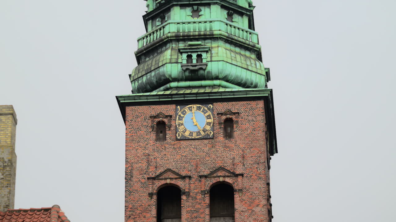 Brick tower with a green copper spire and a blue clock face against a cloudy sky. Copenhagen, Denmark