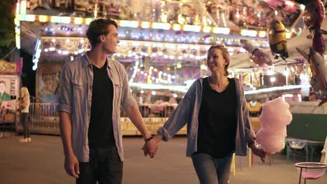 beautiful young couple visiting funfair, smiling, holding hands walking and eating cotton candy, night outdoors. man and woman enjoying day out activities, recreation leisure lifestyle