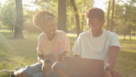 african american friends chatting, having coffee and using laptop in park