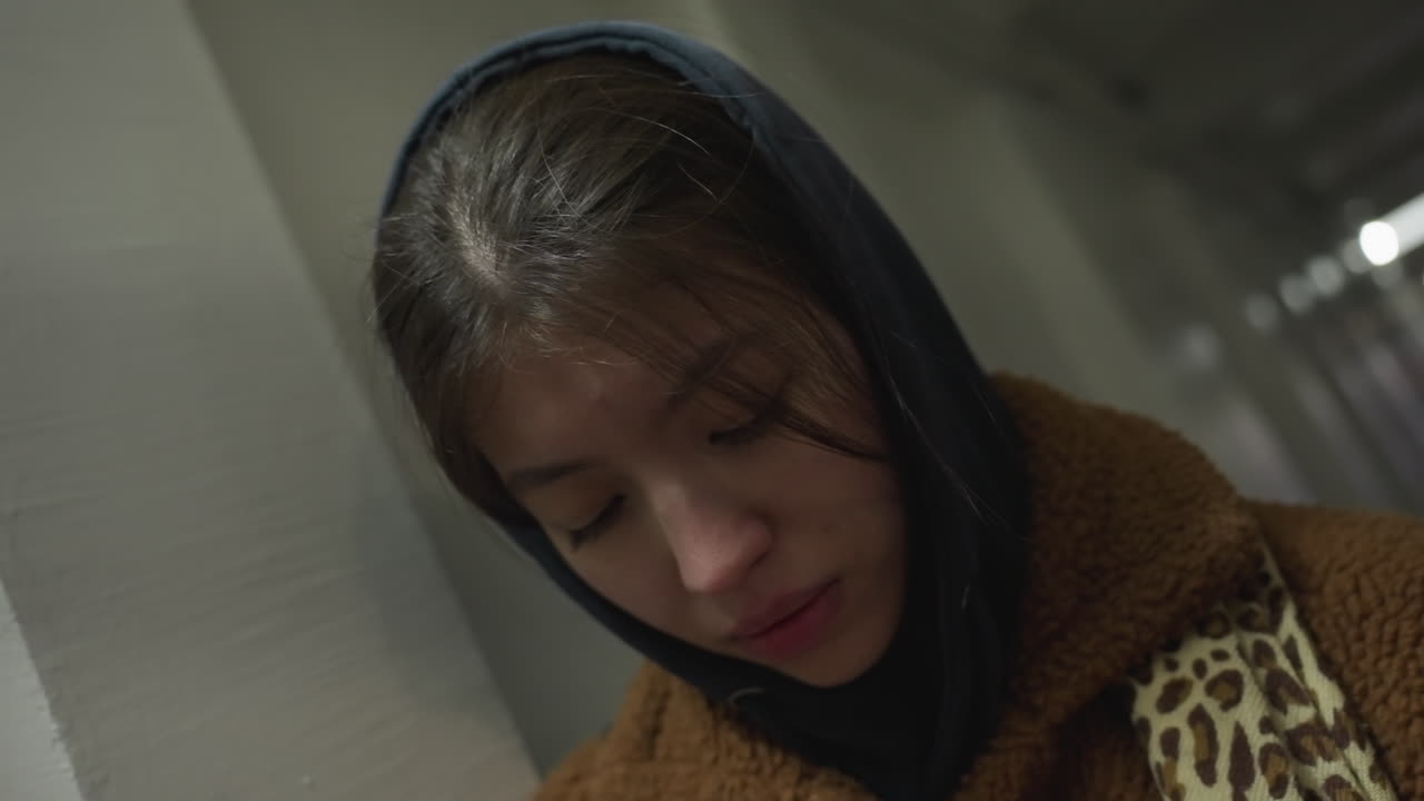 갈색 코트를 입은 곤란한 소녀가 지하 통로의 벽에 기대어있는 클로즈업. 그녀의 가라앉은 눈과 우울한 표정은 희미하게 밝은 환경에서 깊은 감정적 고통을 전달합니다.