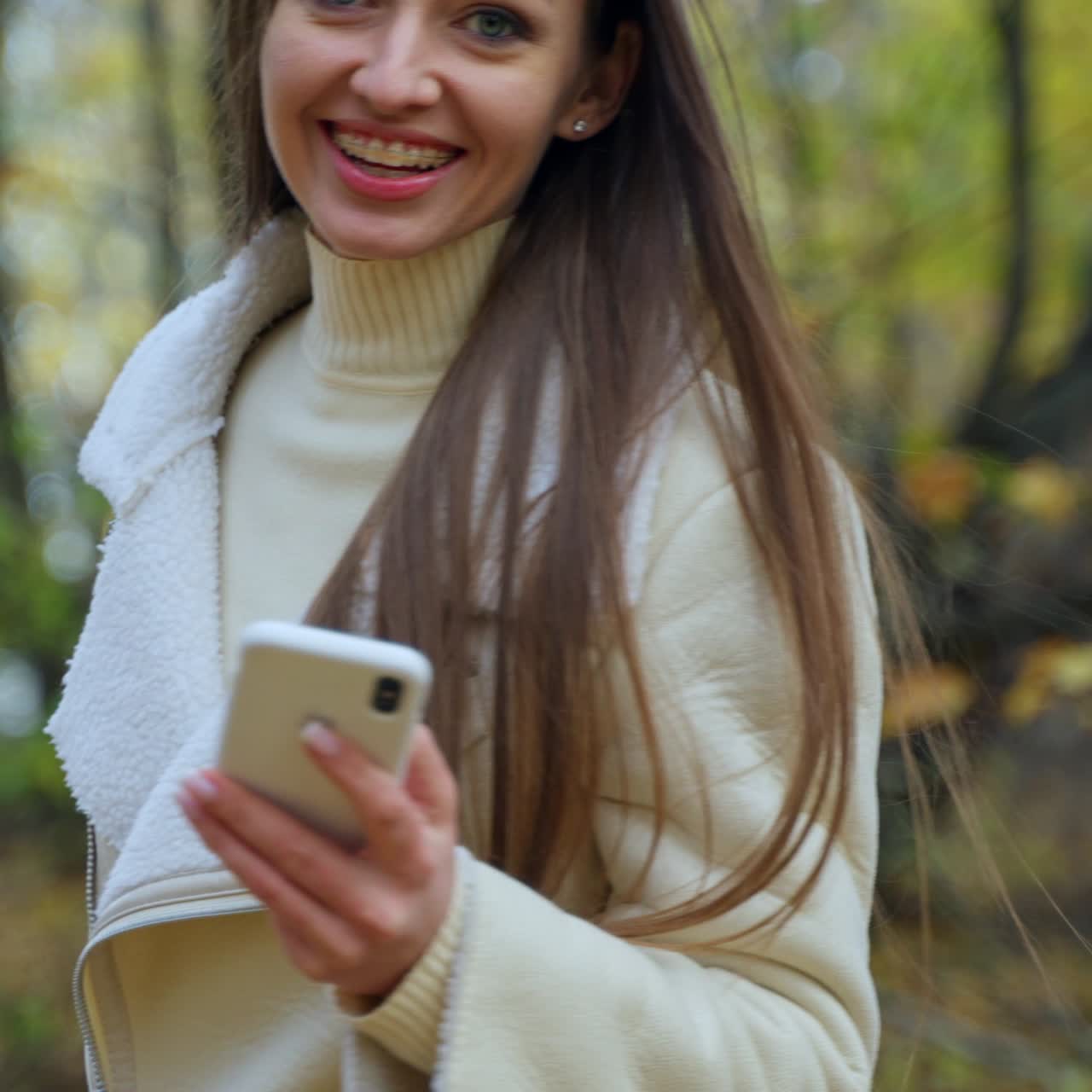 Un'attraente bruna cammina nella natura autunnale con il telefono in mano. La signora si allontana dalla fotocamera, si gira un momento sorridendo e si volta di nuovo.