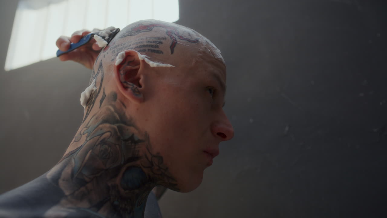 Tattooed Inmate Shaving Head with Razor during Day in Prison Cell