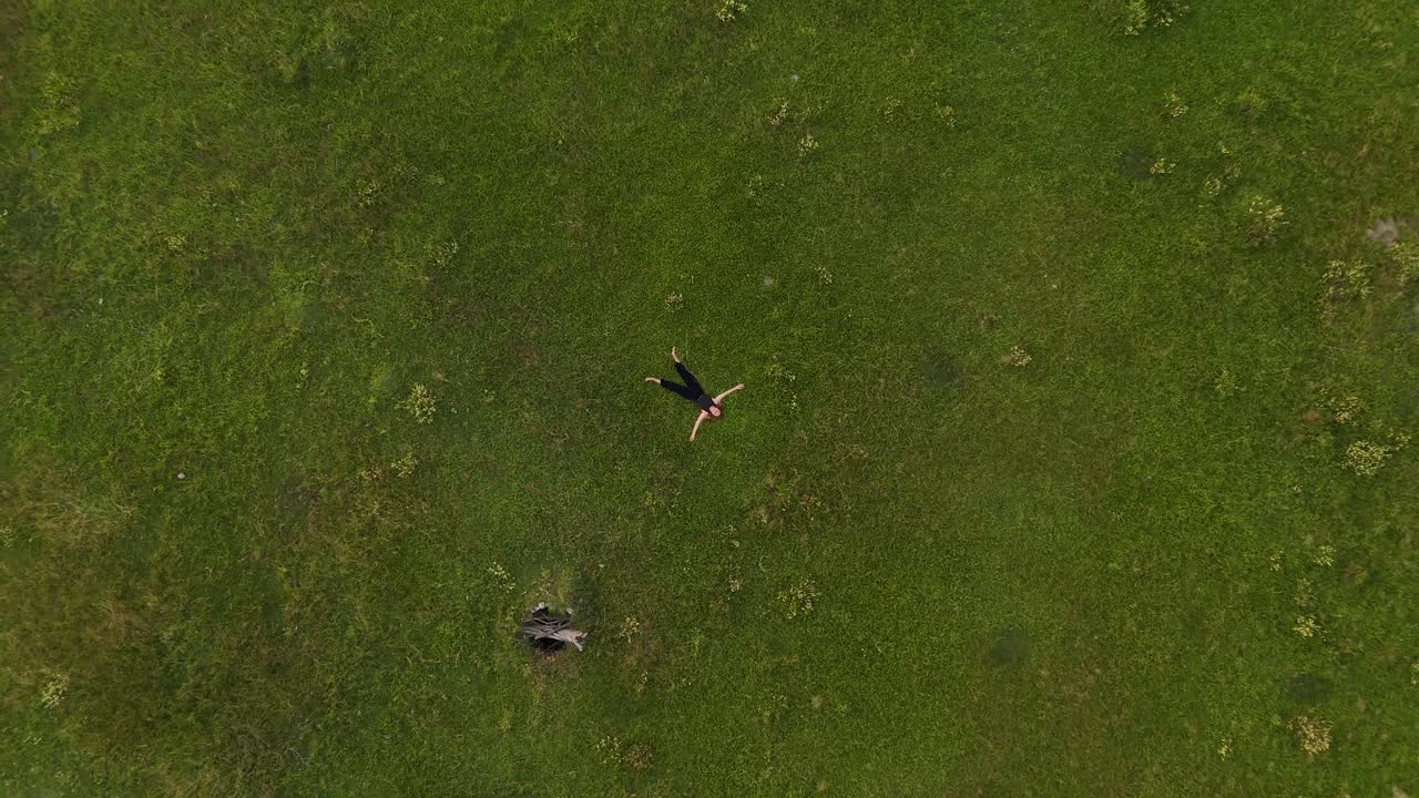 Rising aerial top-down view of a serene young woman lying peacefully on open grassy land, gazing up at the sky.