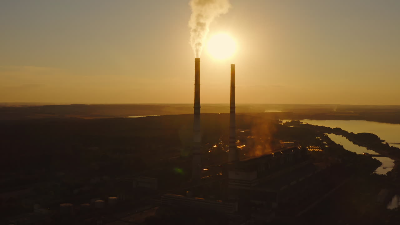 Factory pollutes the atmosphere with harmful emissions. Silhouette of industrial plant among nature. Smoke goes from pipes at sunset.