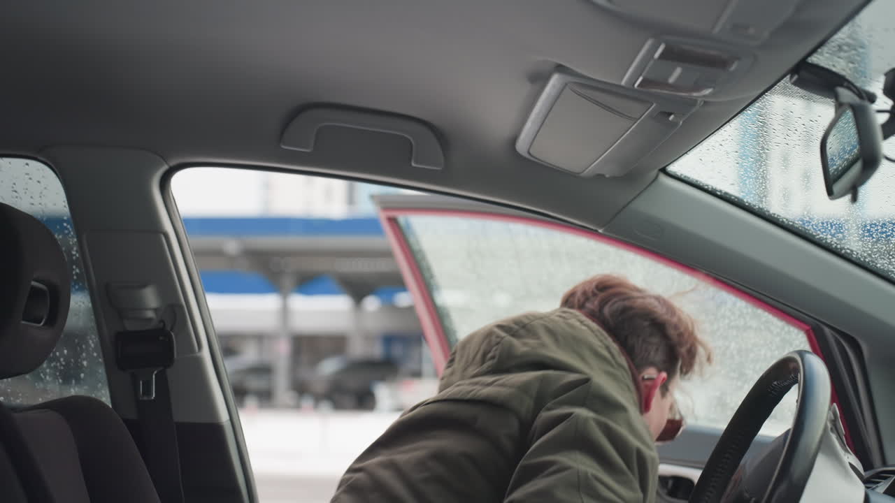 Young boy in winter jacket exits car during cold season with water droplets on window, urban background softly blurred through glass, interior details visible including seat and roof handle