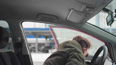 young boy in winter jacket exits car during cold season with water droplets on window, urban background softly blurred through glass, interior details visible including seat and roof handle