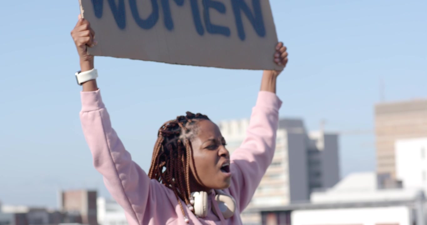 Eine erwachsene Frau beginnt, ein Schild hochzuheben, das protestierende Frauen zeigt, auf einem Dach, ein pinkes Oberteil und ein Headset tragend.