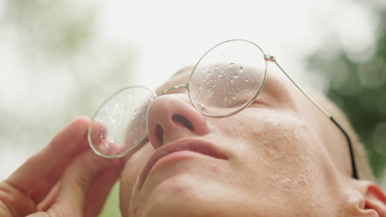 젊은 남자가 젖은 안경을 들어 올려 시야를 확보하고, 둥근 프레임에서 빗방울이 흘러내리는 가운데, 차분한 숨결과 함께 흐린 하늘을 올려다보며 안도하는 표정으로 야외에서 촬영한 인물 사진. 자연 배경과 새롭게 집중된 시선이 돋보입니다.