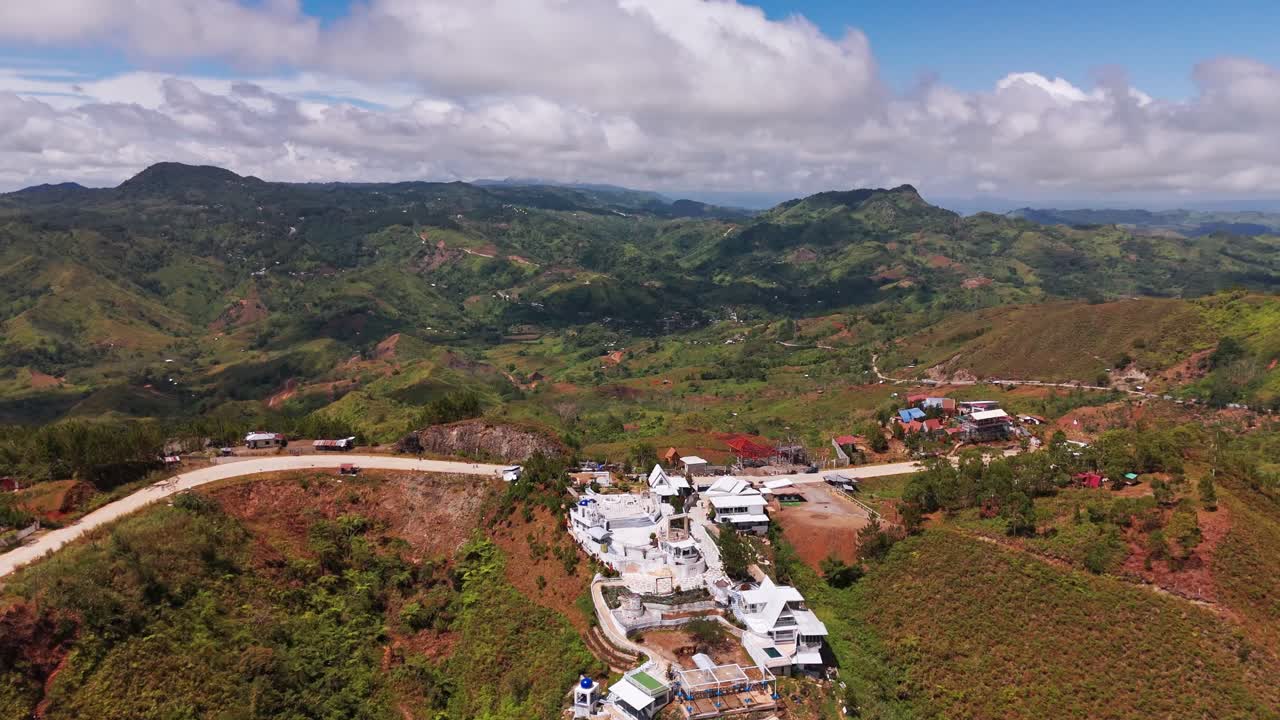 Aerial 4K shot of a remote mountain village surrounded by rolling green hills, winding roads, and dramatic cloudy sky. Scenic highland landscape captured in daylight