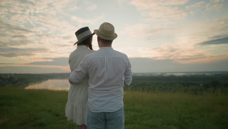 close-up of lovers standing on a grassy hill at sunset, gazing at the sky. the man, dressed in a white shirt, hat, and jeans, gently holds the woman by the waist. she wears a hat and a white dress