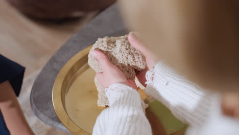 high angle of mindful guest gently lifting and smoothing soft wet soil mold with both hands during calming sensory therapy session, showing tactile interaction on golden tray