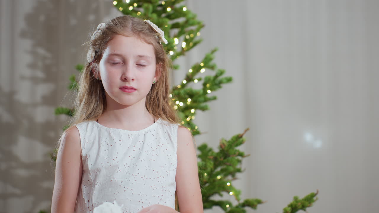 Young girl in white gown standing calmly in front of decorated christmas tree with soft lights, wearing white flower hair accessory, creating peaceful and festive atmosphere during holiday season